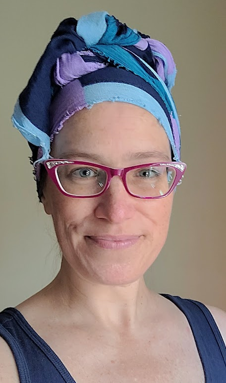 A Caucasian middle-aged woman smiling, wearing purple glasses, a navy blue tank top, and a colorful head wrap.